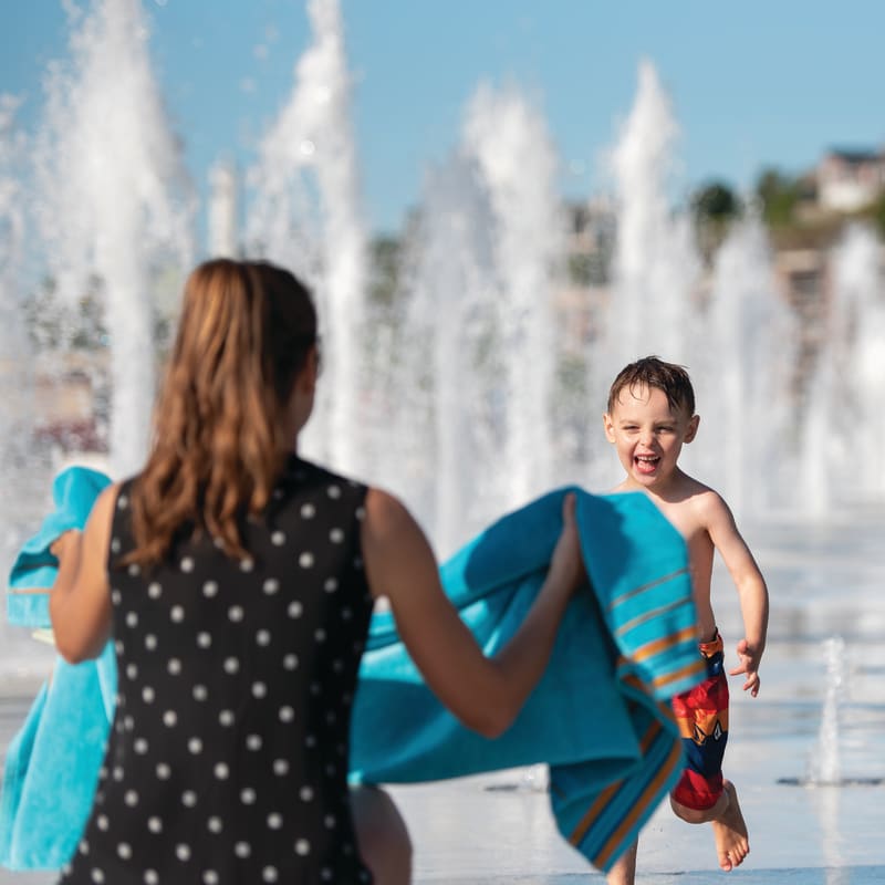 Young boy in water playground happily running towards his mother, who is holding a towel.