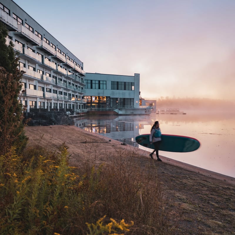 A woman holding a paddleboard at the edge of a lake at dawn.
