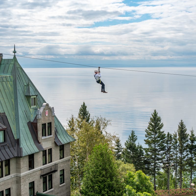 A person on a zipline above the Fairmont Le Manoir Richelieu, with the St. Lawrence River in the background.