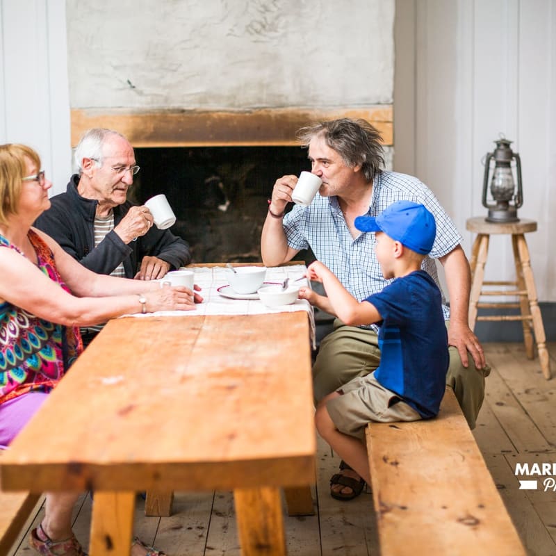 Three adults and a child are sitting on benches around a wooden table in a room with period décor.