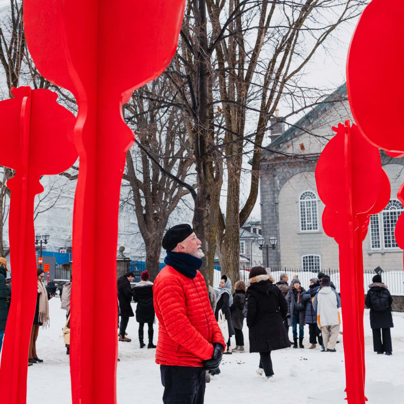 Man looking at an imposing art installation, the red of his coat and the artwork contrasting with the winter landscape.