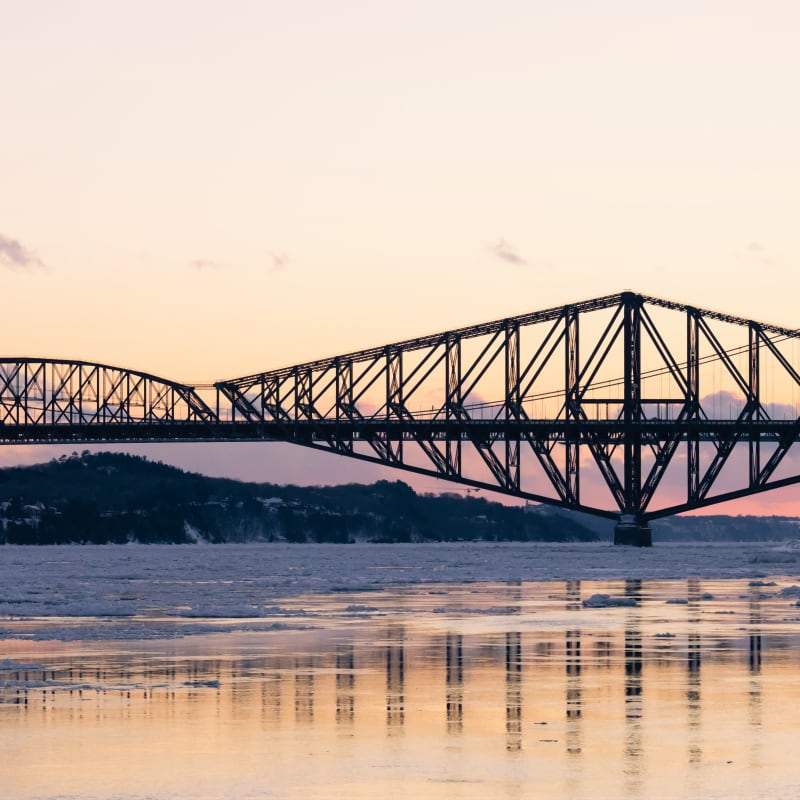 A person by the St. Lawrence River at sunset, with the Pont de Québec bridge on the horizon.