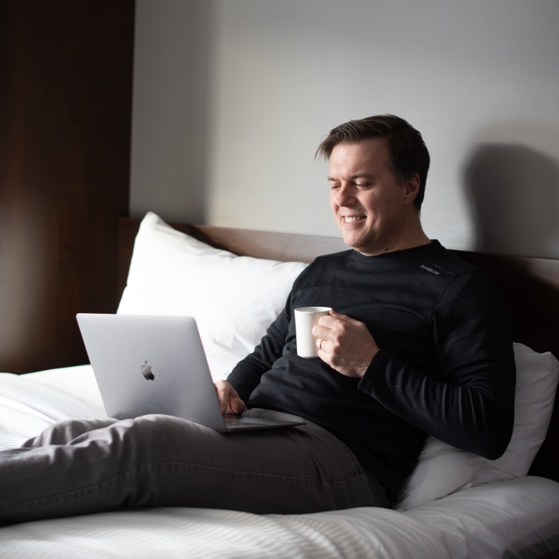Homme allongé sur un lit, avec un café en main et un ordinateur portable sur ses cuisses.