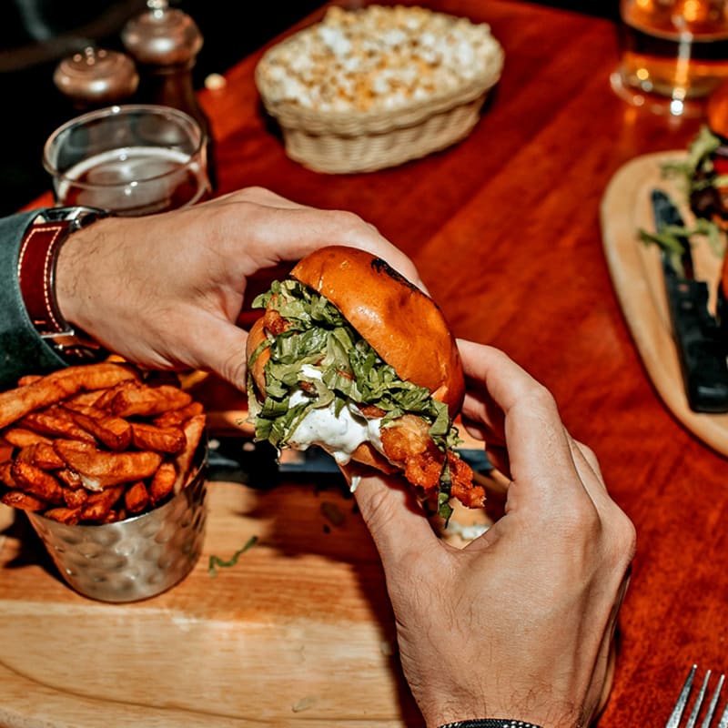 Two hands holding a hamburger above a board with fries on a restaurant table.
