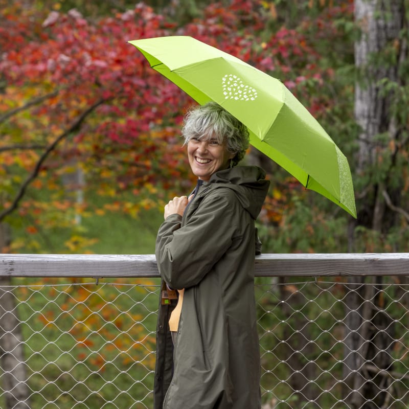 Femme souriante qui s'abrite sous un parapluie vert, avec un paysage automnal en arrière-plan.