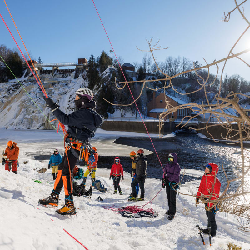 A person is about to start climbing an icy wall on a sunny day; behind them is a group of spectators.