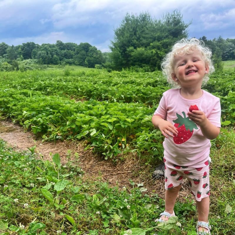 A young child wearing strawberry-patterned clothes eating a strawberry in a field.